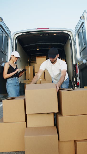 Two movers from Man and Van Epsom are busy loading and packing cardboard boxes into the back of a van, which is parked on a paved area near a residential property. The man, wearing a black cap and white shirt, is actively lifting a box and placing it inside the vehicle, while a woman, dressed in a white cap, black top, and jeans, is standing nearby holding a clipboard, possibly checking items or coordinating the packing process. The open rear doors of the van reveal additional packed boxes stacked inside, ready for a home relocation or furniture transport. Various packing materials, such as plastic-wrapped items and cardboard boxes, are visible, indicating an organized moving and packing operation. The environment is well-lit by natural daylight, and the scene depicts a professional moving service preparing for a house move, supporting efficient loading and logistical planning for customers near Epsom.