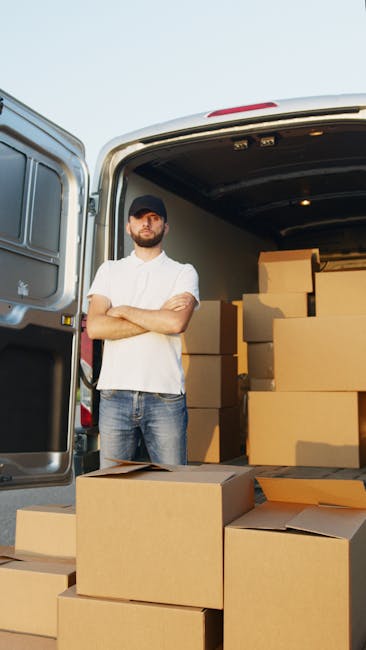 A man wearing a black cap, white shirt, and jeans stands with arms crossed beside an open van parked outdoors, ready for a home relocation. The van's interior shows neatly stacked cardboard boxes, some open with packing paper visible, indicating an ongoing packing and moving process. The boxes vary in size and are arranged on the pavement, with additional boxes placed on the ground nearby. The scene is illuminated by natural daylight, and the backdrop includes a clear sky, suggesting a calm and organized furniture transport operation managed by Man and Van Epsom. This image captures the preparation stage of professional removals, emphasizing careful packing and loading involved in house removals near Epsom Station, EPSOM.