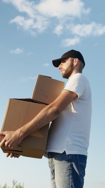 Two movers from Man and Van Epsom are busy loading and packing cardboard boxes into the back of a van, which is parked on a paved area near a residential property. The man, wearing a black cap and white shirt, is actively lifting a box and placing it inside the vehicle, while a woman, dressed in a white cap, black top, and jeans, is standing nearby holding a clipboard, possibly checking items or coordinating the packing process. The open rear doors of the van reveal additional packed boxes stacked inside, ready for a home relocation or furniture transport. Various packing materials, such as plastic-wrapped items and cardboard boxes, are visible, indicating an organized moving and packing operation. The environment is well-lit by natural daylight, and the scene depicts a professional moving service preparing for a house move, supporting efficient loading and logistical planning for customers near Epsom.