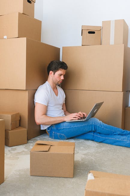 A young man with dark hair, wearing a white T-shirt and blue jeans, sitting on a concrete floor surrounded by numerous cardboard boxes of varying sizes, some sealed with tape and others open, in an indoor space with plain white walls. He is working on a silver laptop, with one hand on the keyboard, during a home relocation process. Large cardboard boxes are stacked against the wall behind him, indicating a packing phase for a house move, which may involve furniture transport and logistical planning. The area appears to be a room or storage space prepared for the moving day, with no furniture present besides the boxes, and the lighting is neutral and even, suggesting daytime or well-lit conditions. This scene illustrates the packing and loading preparations typical of professional removals, such as those offered by Man and Van Epsom, for house or flat moves from Ashley Road to KT17, reflecting the process of organising belongings for transportation.