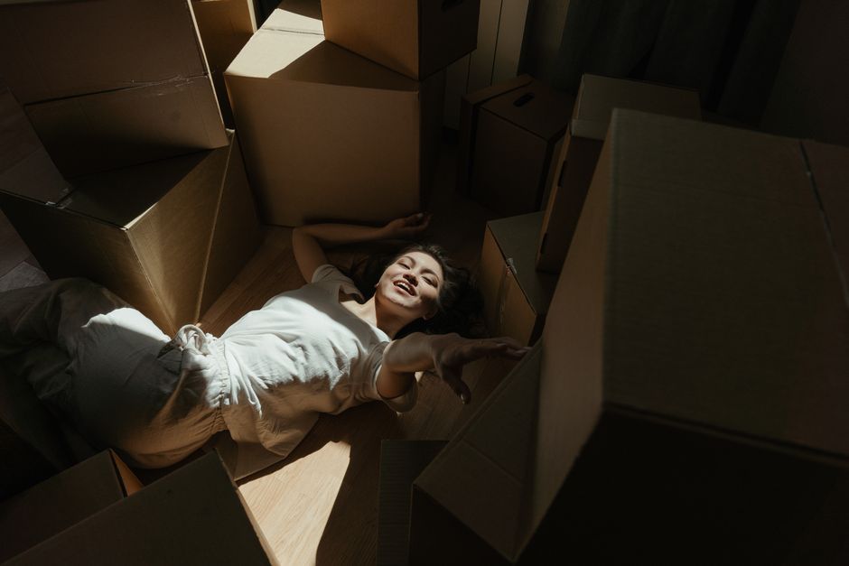 A woman with dark hair, wearing a white shirt and light-colored pants, is lying on the wooden floor inside a room surrounded by cardboard moving boxes of various sizes. Some boxes are stacked, while others are scattered around her. She appears to be smiling and relaxed, with her head resting on one of the boxes. The boxes are made of brown corrugated cardboard, with some wrapped in plastic or packing materials visible. The room is dimly lit, with natural light casting shadows on the boxes and the woman. The scene depicts the process of home relocation, with the woman enjoying a break amid packing and moving preparations, related to the services offered by Man and Van Epsom for house removals and furniture transport.