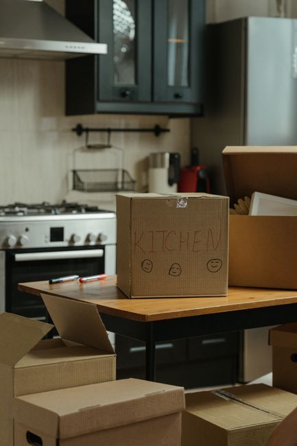 A young man with dark hair, wearing a white T-shirt and blue jeans, sitting on a concrete floor surrounded by numerous cardboard boxes of varying sizes, some sealed with tape and others open, in an indoor space with plain white walls. He is working on a silver laptop, with one hand on the keyboard, during a home relocation process. Large cardboard boxes are stacked against the wall behind him, indicating a packing phase for a house move, which may involve furniture transport and logistical planning. The area appears to be a room or storage space prepared for the moving day, with no furniture present besides the boxes, and the lighting is neutral and even, suggesting daytime or well-lit conditions. This scene illustrates the packing and loading preparations typical of professional removals, such as those offered by Man and Van Epsom, for house or flat moves from Ashley Road to KT17, reflecting the process of organising belongings for transportation.