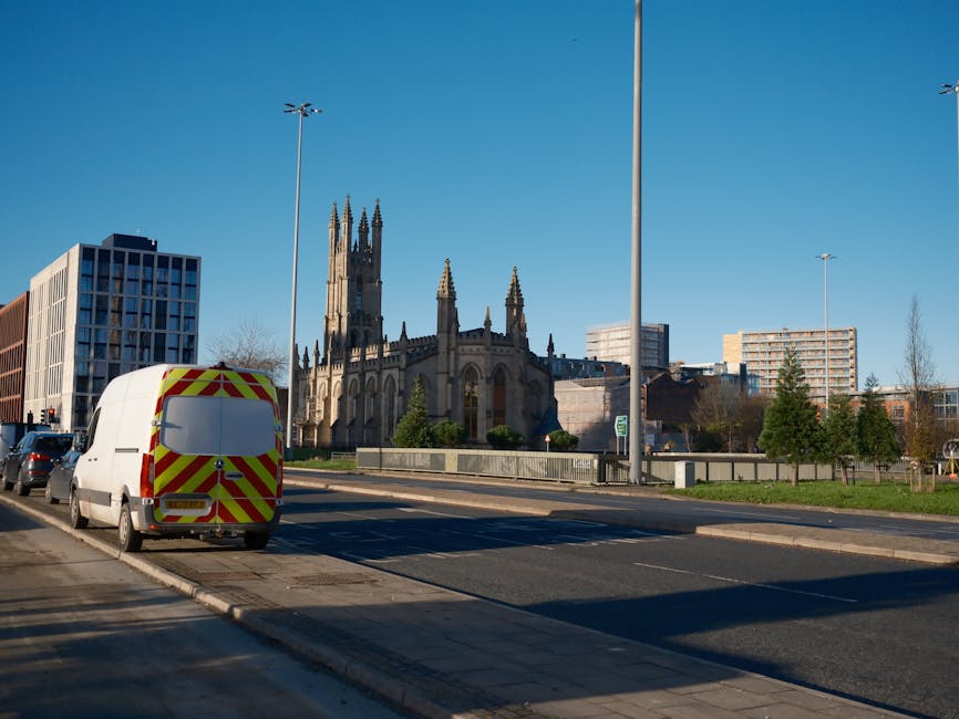 A white cargo van with reflective red and yellow chevron markings on its rear doors is parked on the side of a city street during daylight. The van belongs to Man and Van Epsom, a professional removals service, and is positioned near a pedestrian pavement, adjacent to a church with tall, Gothic-style architecture featuring pointed spires and arched windows. In the background, modern office buildings and residential flats are visible under a clear blue sky. The scene includes street lamps, a few trees, and some parked cars along the road. The van appears to be in the process of either loading or unloading goods for a home relocation or furniture transport, emphasizing the logistics involved in packing, moving, and transportation services offered by the company. The environment is well-lit with natural sunlight, highlighting the urban surroundings and the vehicle’s placement for a moving operation.