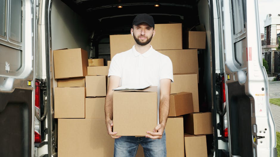 A man wearing a white polo shirt, dark cap, and jeans stands at the open rear of a white delivery van, holding a cardboard box during a house removal or relocation process. The van's interior is filled with stacked cardboard boxes of various sizes, some secured with plastic wrapping, and selected items are protected with moving blankets. The man appears to be preparing to load or unload the boxes as part of furniture transport or packing and moving activities. Light from outside illuminates the scene, revealing a residential street with houses and greenery in the background. This image captures the logistics involved in house removals, facilitated by the services of Man and Van Epsom.