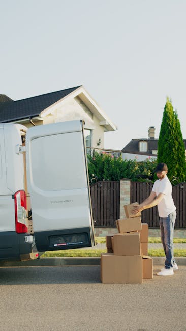 A white cargo van with reflective red and yellow chevron markings on its rear doors is parked on the side of a city street during daylight. The van belongs to Man and Van Epsom, a professional removals service, and is positioned near a pedestrian pavement, adjacent to a church with tall, Gothic-style architecture featuring pointed spires and arched windows. In the background, modern office buildings and residential flats are visible under a clear blue sky. The scene includes street lamps, a few trees, and some parked cars along the road. The van appears to be in the process of either loading or unloading goods for a home relocation or furniture transport, emphasizing the logistics involved in packing, moving, and transportation services offered by the company. The environment is well-lit with natural sunlight, highlighting the urban surroundings and the vehicle’s placement for a moving operation.