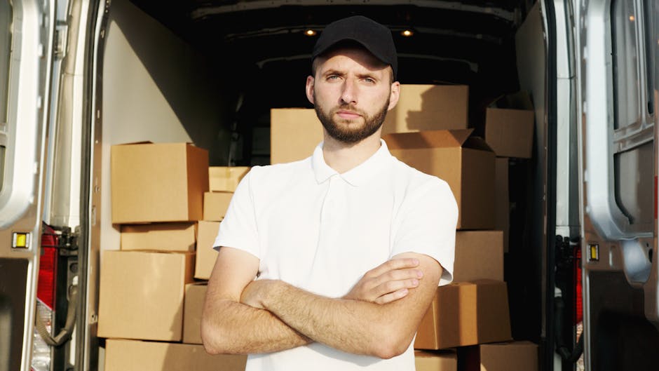 A man wearing a black cap, black vest, and blue long-sleeve shirt is standing at the rear of a white commercial van, holding a cardboard box in both hands. The van's open side door reveals several oth